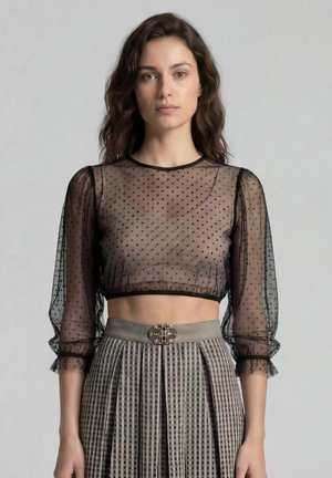 Frau mit welligem braunem Haar, die ein durchsichtiges schwarzes bauchfreies Top mit Polka-Dots und einen hoch taillierten grauen plissierten Rock mit dekorativer Gürtelschnalle trägt.