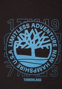 Zwarte T-shirt met een blauw circulair grafisch ontwerp van een boom en de tekst "LIMITLESS ADVENTURE NEW HAMPSHIRE U.S.A TIMBERLAND."