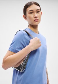 Svetlo modra majica s kratkimi rokavi in okroglim izrezom. Model drži srebrno kovinsko torbico z gladko teksturo in elegantnim dizajnom.