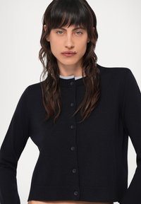 Ναυτικό μπλε πλεκτό κασκόλ με στρογγυλή λαιμόκοψη, κλείσιμο με κουμπί και μακριά μανίκια. Λείος υφή και κοντή σχεδίαση.