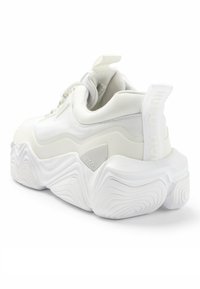 Bílé tenisky s texturovanou síťovinou a hladkými překrytími, se silnou vlnitou podrážkou a značkovým detailem na patním pásku.