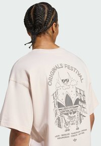 Oversized ljusrosa bomulls-t-shirt med en grafisk design på baksidan, märkt "Originals Festival", med abstrakta karaktärer och stadsnamn.
