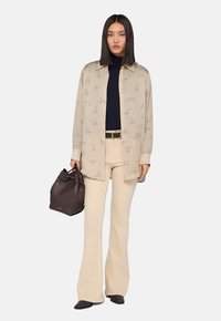 Donna con capelli neri lisci che indossa una camicia button-up beige stampata sopra un top nero, pantaloni a campana crema, mentre tiene una borsa a secchiello in pelle marrone scuro.