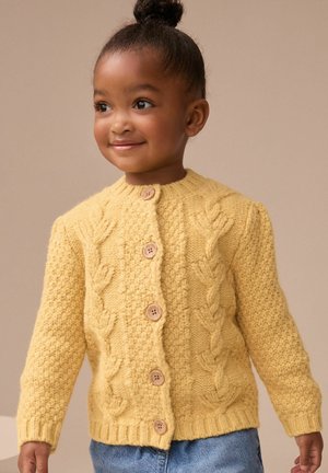 Junges Mädchen mit dunklen Haaren im Dutt, trägt eine gelbe gestrickte Strickjacke mit Knöpfen und blaue Jeans, lächelt und schaut zur Seite.