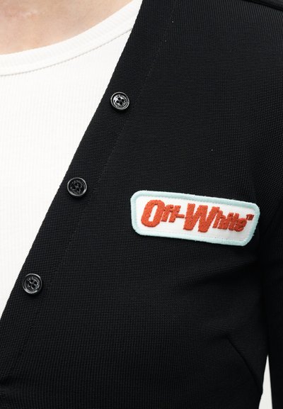 Cardigan noir boutonné porté sur une chemise blanche avec un patch rectangulaire du logo Off-White cousu sur le côté gauche de la poitrine.