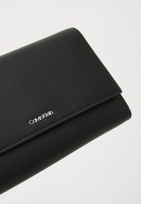 Black leather wallet with a textured finish, featuring a silver logo and sleek design. Fold-over closure with visible stitching detail.