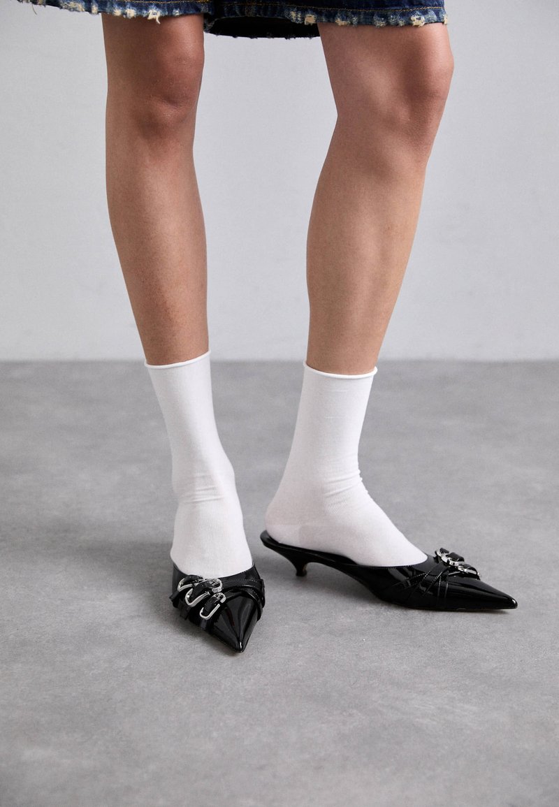 Mulas de charol negro con puntas afiladas, tacones bajos y herrajes decorativos de plata, combinadas con calcetines blancos hasta el tobillo.
