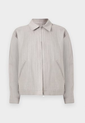 Muž oblečený v béžovom obleku so širokými nohavicami, zodpovedajúca bunda na zips, čierna košeľa a čierne topánky, stojaci na bielom pozadí.