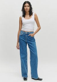 Blauwe wijde spijkerbroek met knoop- en ritssluiting, vijf zakken en een klassieke denimtextuur, gecombineerd met een witte aansluitende tanktop.