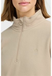 Beige fleecjacket met hoge kraag; heeft een gedeeltelijke rits en een subtiele geborduurde logo aan de linkerkant. Gladde textuur.
