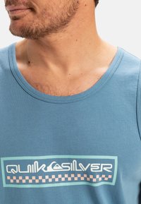 Blå bomulds tanktop med et grafisk print i hvid og lyseblå med et ternet kantbånd i pink. Rund halsudskæring og uden ærmer.