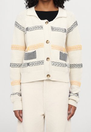 Cardigan couleur crème avec un col, orné de rayures horizontales de motifs jaunes, noirs et bleus, et de gros boutons à l'avant.