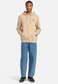 Beige hoodie with kangaroo pocket and drawstring hood, paired with light blue denim jeans, and black sneakers. Logo detail on the chest.