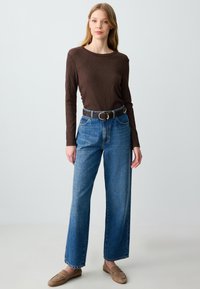 Bruine lange mouw geribbelde top, hooggeknoopte lichtblauwe jeans, donkere riem met ronde gesp en beige instappers. Eenvoudig recht ontwerp.
