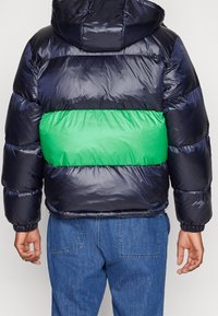 Doudoune avec une partie supérieure marine et une bande horizontale vert vif au milieu. Finition brillante avec poignets élastiques.
