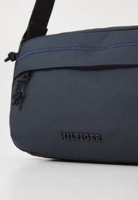 Námorníckomodrá crossbody taška z odolnej látky, šikmá zipsová kapsa a textúrované logo. Má nastaviteľný popruh a jedinečné šnúrkové akcenty.