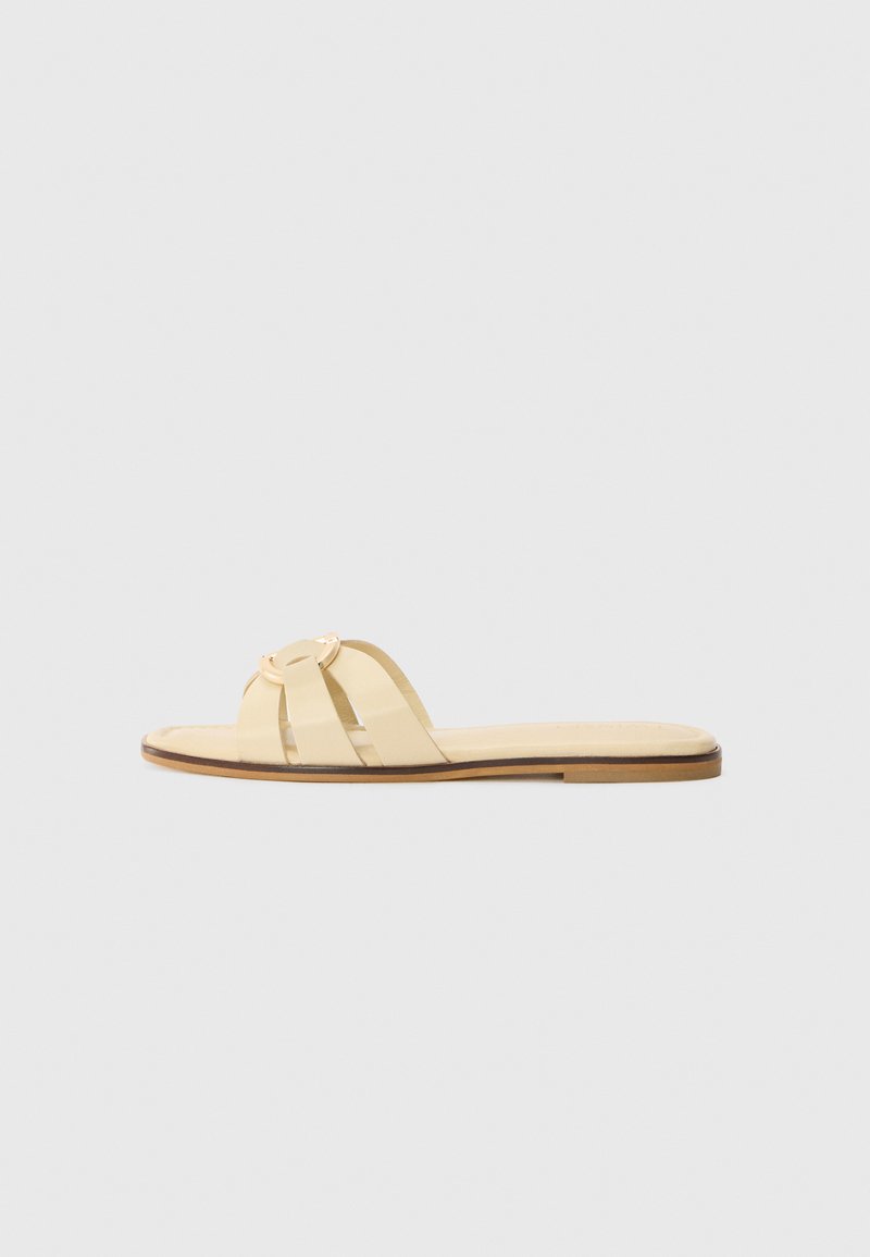 Flat sandal made of pale yellow leather, featuring two wide straps with a circular gold accent. The sole is a light brown.