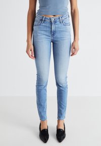 Jeans skinny bleu clair, taille haute, avec une texture lisse, cinq poches et un léger délavage. Associés à des chaussures noires à bout pointu.