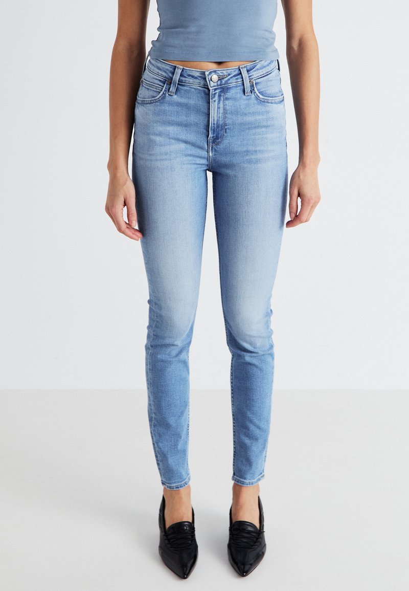 Jeans skinny bleu clair, taille haute, avec une texture lisse, cinq poches et un léger délavage. Associés à des chaussures noires à bout pointu.