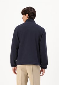 Jachetă din fleece bleumarin cu guler înalt, croială lejeră și mâneci lungi. Materialul pare moale și texturat, potrivit pentru ținute casual.