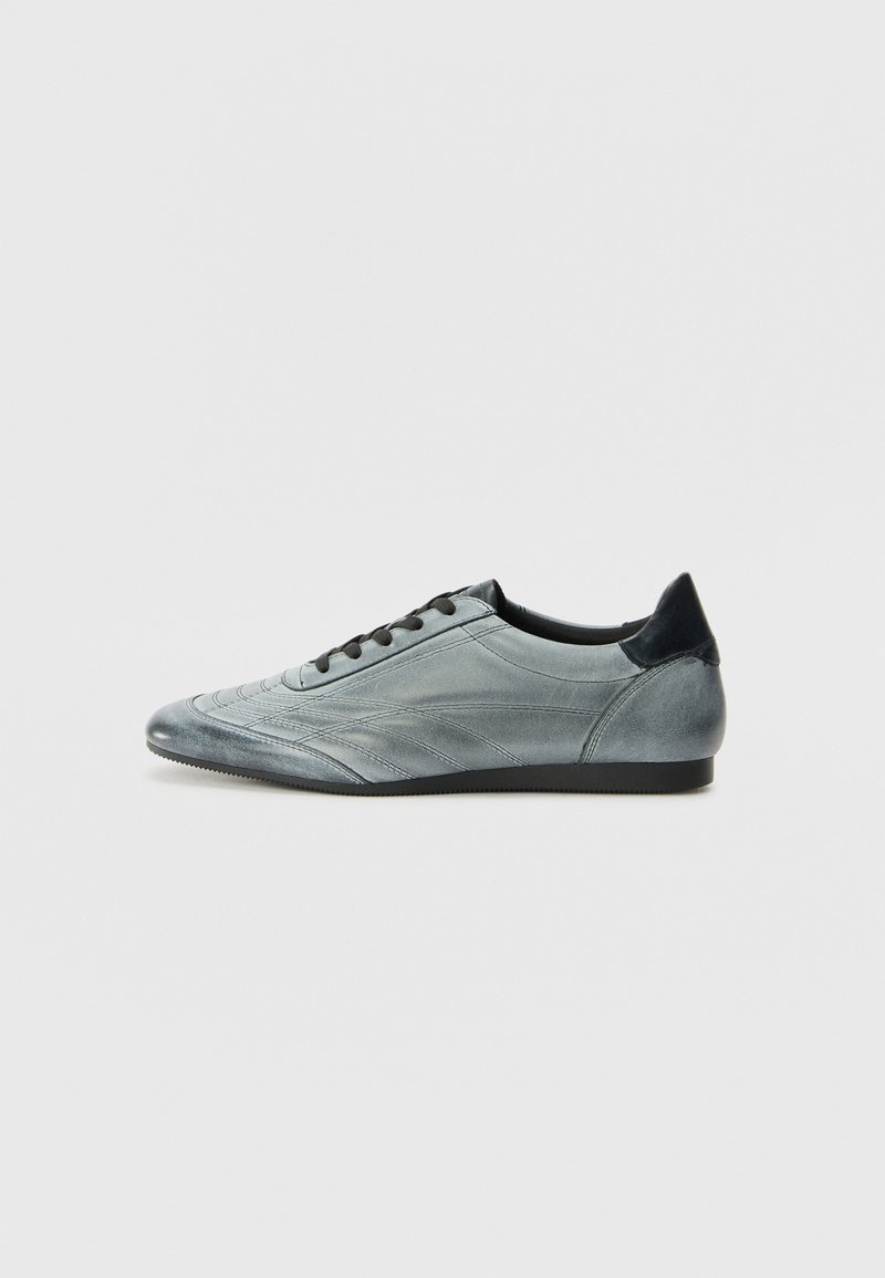 Baskets grises pour homme à profil bas avec lacets noirs et languette de talon noire, présentées sur un fond gris clair uni.