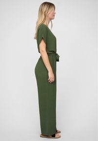 Woman standing sideways wearing a long olive green dress with a waist tie and black sandals on a gray background.