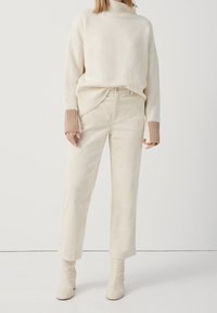 Krämfärgad chunky stickad turtlenecktröja med ribbade ärmslut, i kombination med krämfärgade manchesterbyxor med vida ben och ankelskor.