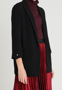 Žena v černém šitém saku přes vínový rolák a červenou plisovanou sukni na jednobarevném pozadí.