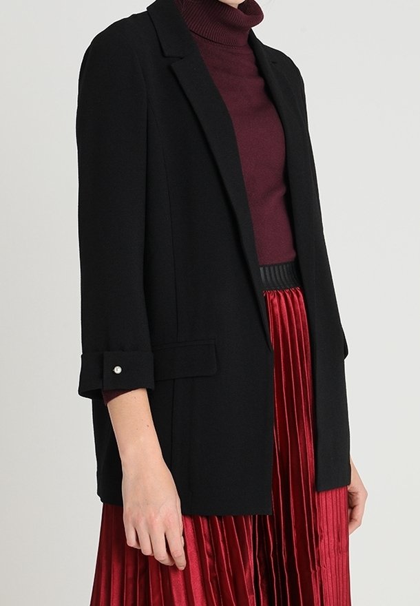 Žena v černém šitém saku přes vínový rolák a červenou plisovanou sukni na jednobarevném pozadí.