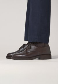 Braune Leder Schuhe mit strukturiertem Finish, Schnürdesign und Gummisohle, kombiniert mit dunklen Socken und Hosen.