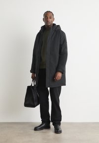 Schwarze wasserdichte Parka mit Kapuze, vorn mit Reißverschluss, schlanker Schnitt und weicher Textur. Kombiniert mit dunklen Hosen und einer schwarzen Tragetasche.