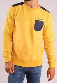 Felpa gialla con maniche testurizzate e una tasca blu scuro. Polsini e fondo a costine, con un logo sul petto. Fit e design casual.