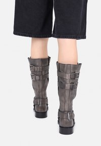 Persona que lleva botas de cuero gris oscuro hasta la mitad de la pantorrilla con tres correas ajustables y hebillas, de pie frente a un fondo blanco.