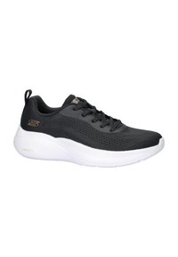 Zapatilla deportiva negra con parte superior de malla texturizada, diseño con cordones, suela blanca acolchada y sutil marca dorada.