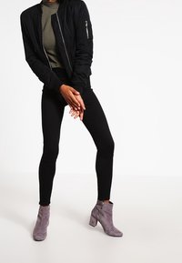 Veste bombardier noire, top vert olive, jean skinny noir, bottines en daim gris avec talon épais et détail zippé.