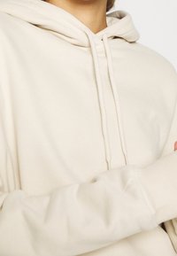 Sudadera beige con una textura suave, capucha con cordón y mangas largas con puños de canalé. Diseño sencillo sin patrones ni acentos visibles.