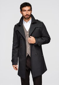 Homme portant un trench-coat noir croisé sur un gilet beige et une chemise blanche, ajustant le manteau de la main droite, regardant droit devant.
