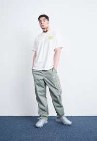 Ανδρική εμφάνιση με κρεμ μπλουζάκι με πράσινο γραφικό και ελαφρώς φαρδιά, ανοιχτό πράσινα cargo παντελόνια. Γκρι αθλητικά παπούτσι�α ολοκληρώνουν την εμφάνιση.