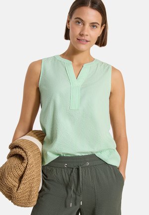 Femme portant un haut sans manches rayé vert menthe et un pantalon vert foncé à cordon, tenant un tapis tissé roulé dans une main.