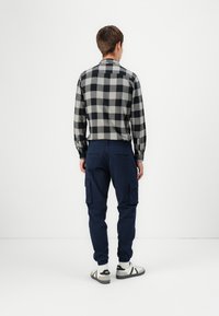 Šedá a černá kostkovaná košile, námořnické cargo kalhoty, bílé boty s černými detaily. Viděno zezadu, zápěstí viditelné, minimalistické pozadí.