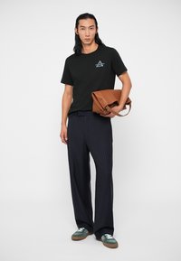 Sort bomuldstrøje med et lille blåt og hvidt grafik, kombineret med navy stribede bukser og grønne sneakers. Brun lædertaske i hånden.