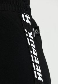 Jogger atlético negro con una textura suave y lisa. Presenta un logo blanco impreso en el costado y una pequeña etiqueta de marca negra.