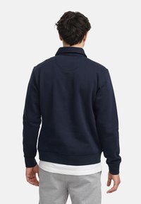 Felpa blu navy con colletto alto, dotata di polsini e orlo a coste, realizzata in un tessuto morbido. Indossata sopra una camicia bianca a strati e pantaloni grigi.