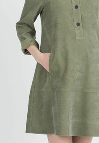 Abito in corduroy verde oliva con maniche a tre quarti, apertura con bottoni e tasche laterali. Tessuto liscio con una leggera lucentezza.