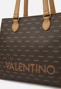 Bolso tote marrón de Valentino con asas beige, con pequeños logotipos de Valentino repetidos y un logotipo grande de Valentino en el frente.