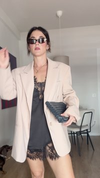 Blazer beige oversize con rever, indossato sopra un top in pizzo nero. Tiene una clutch strutturata blu scuro.