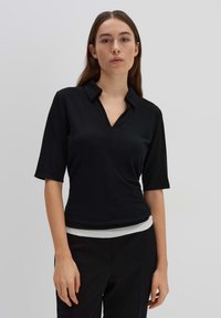 Chemise noire à manches courtes avec col en V, fabriquée en tissu lisse. Portée sur une taille claire. Design minimaliste.