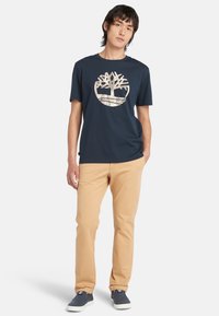 T-shirt in cotone blu navy con logo di un albero beige, abbinato a pantaloni beige chiaro e scarpe slip-on blu navy. Design semplice e casual.