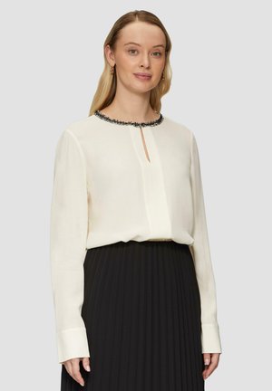 Blouse crème avec un col rond en perles noires, un décolleté en forme de clé à l'avant, des manches longues et une coupe décontractée, associée à une jupe plissée noire.