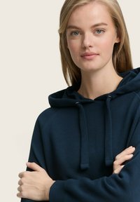 Tamsiai mėlyna kapšelė iš minkštos, tekstūrinės medžiagos su raišteliu ir raglano rankovėmis. Modelis sukryžiuoja rankas, pabrėždamas dizainą.